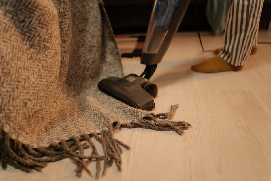 Close-up of a vacuum cleaner performing surface cleaning on a tan, woven rug with frayed edges, placed on a light wooden floor in a domestic setting. The vacuum head is black with a transparent sectio